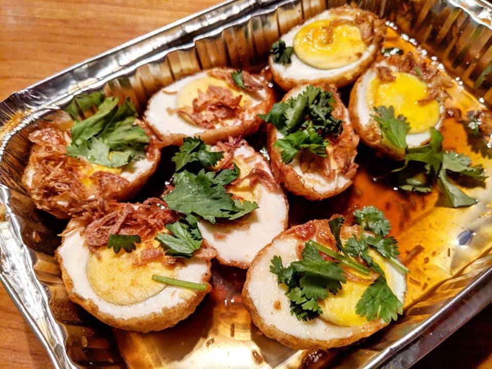 Hard-boiled eggs, sliced in half and fried, sit in an aluminum takeout container, topped with fried shallots, a thin brown sauce, and herbs