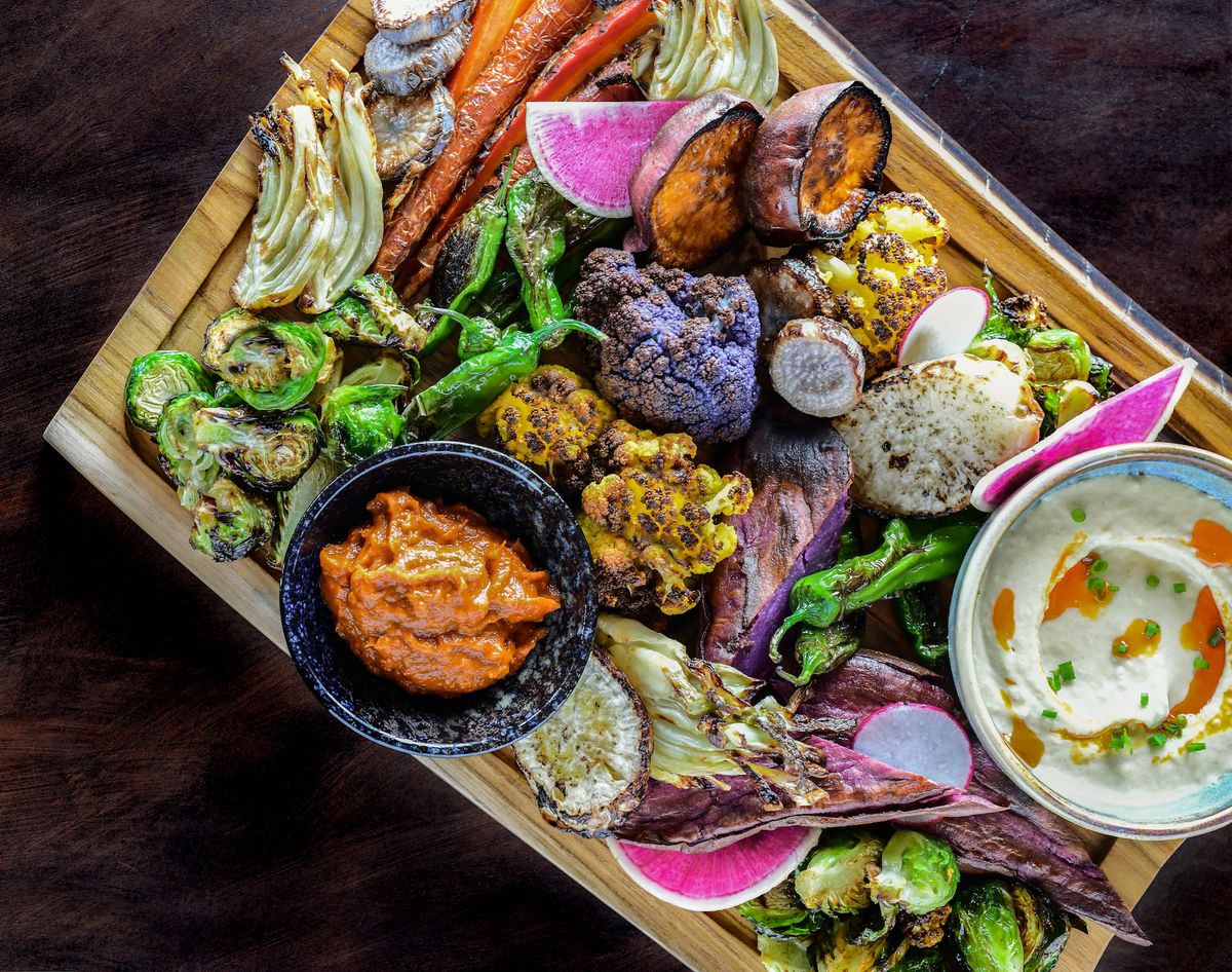 A wooden board piled with vegetables like purple cauliflower, grilled shishito peppers, hummus, watermelon radish, and sweet potato.