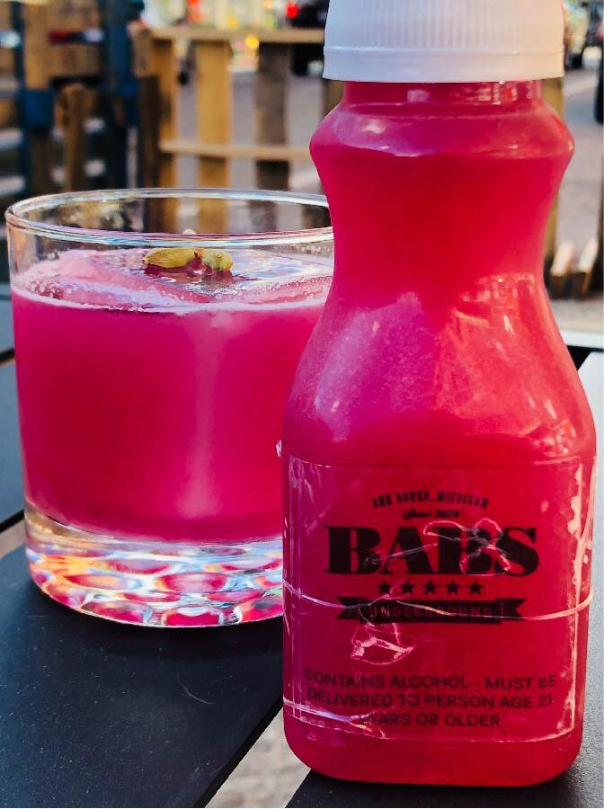 A plastic bottle with a label for Babs is filled with hot pink liquid and sits next to a tumbler of the same cocktail. 