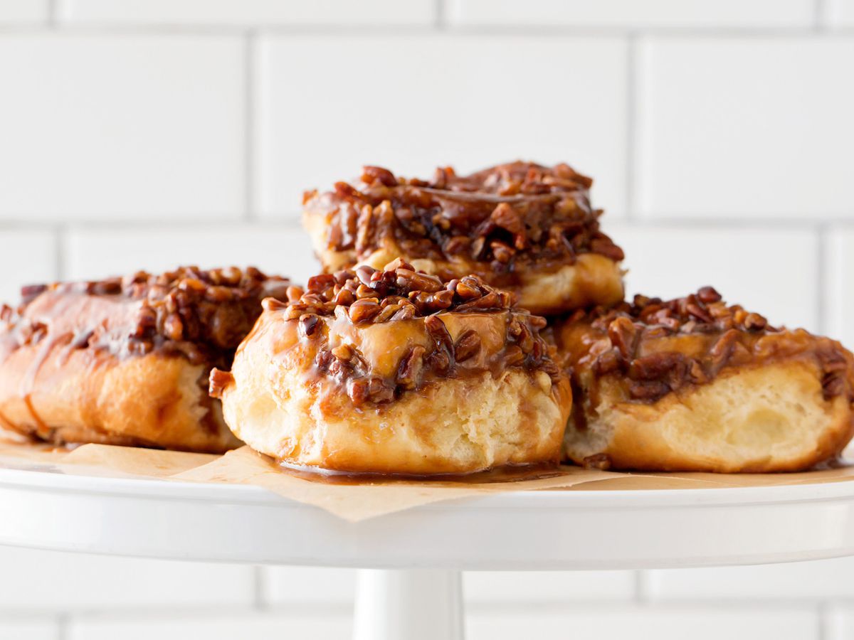 Plump sticky buns, topped with nuts, sit on a white cake display in front of a white tiled wall