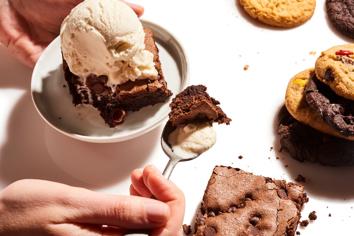Hand holding a spoonful of brownie and vanilla ice cream next to a carton of vanilla ice cream containing a chocolate brownie topped with a scoop of vanilla ice cream. Chocolate chip cookies and brownie are scattered beside the carton from Insomnia Cookies