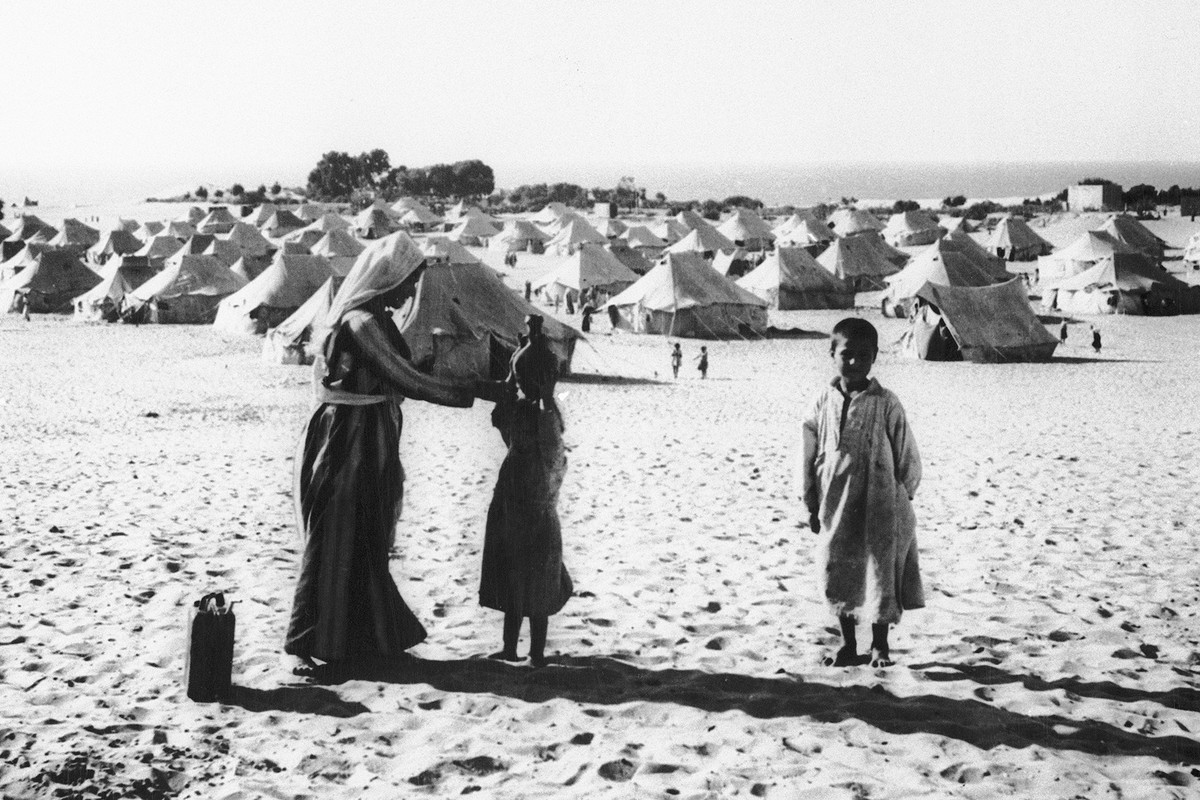 A black-and-white photo shows a woman and two children wearing long tunics standing on the sand. Behind them stretches a tent-city.