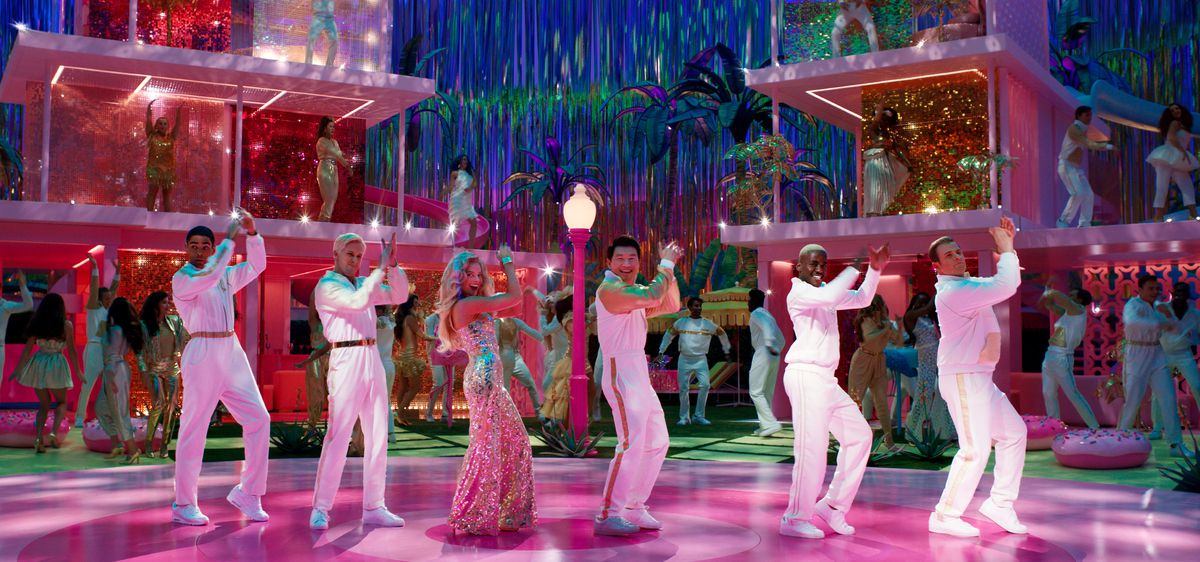 Barbie (Margot Robbie), in a glittery pink gown, does a line dance in front of a pair of wall-less pink plastic life-sized Barbie Dreamhouses, flanked by five Kens in all white, played by Kingsley Ben-Adir, Ryan Gosling, Simu Liu, Ncuti Gatwa, and Scott Evans, in the 2023 movie Barbie