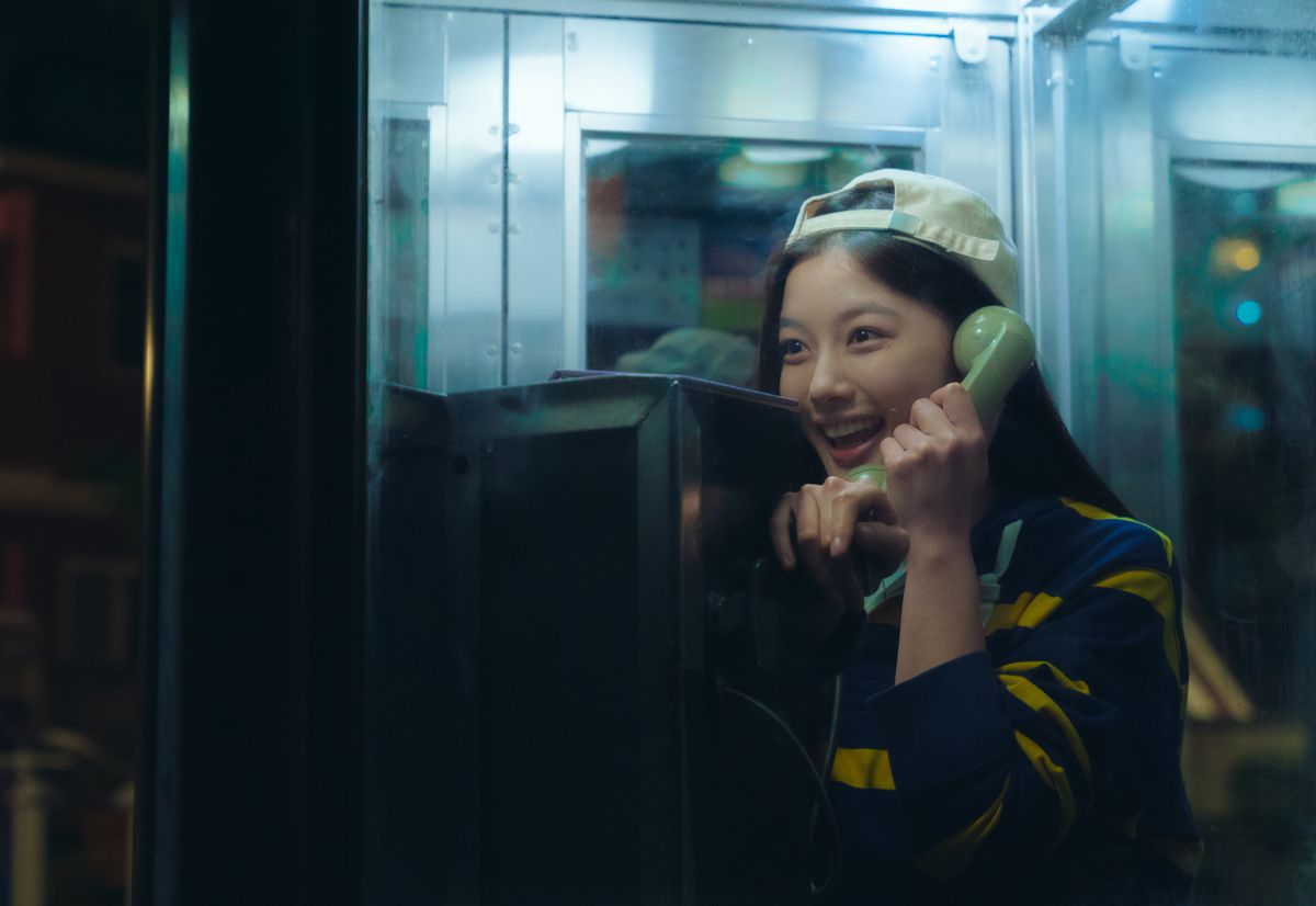A girl wearing a backwards tan baseball cap smiles while talking into the receiving end of a green telephone inside a glass phonebooth at night.