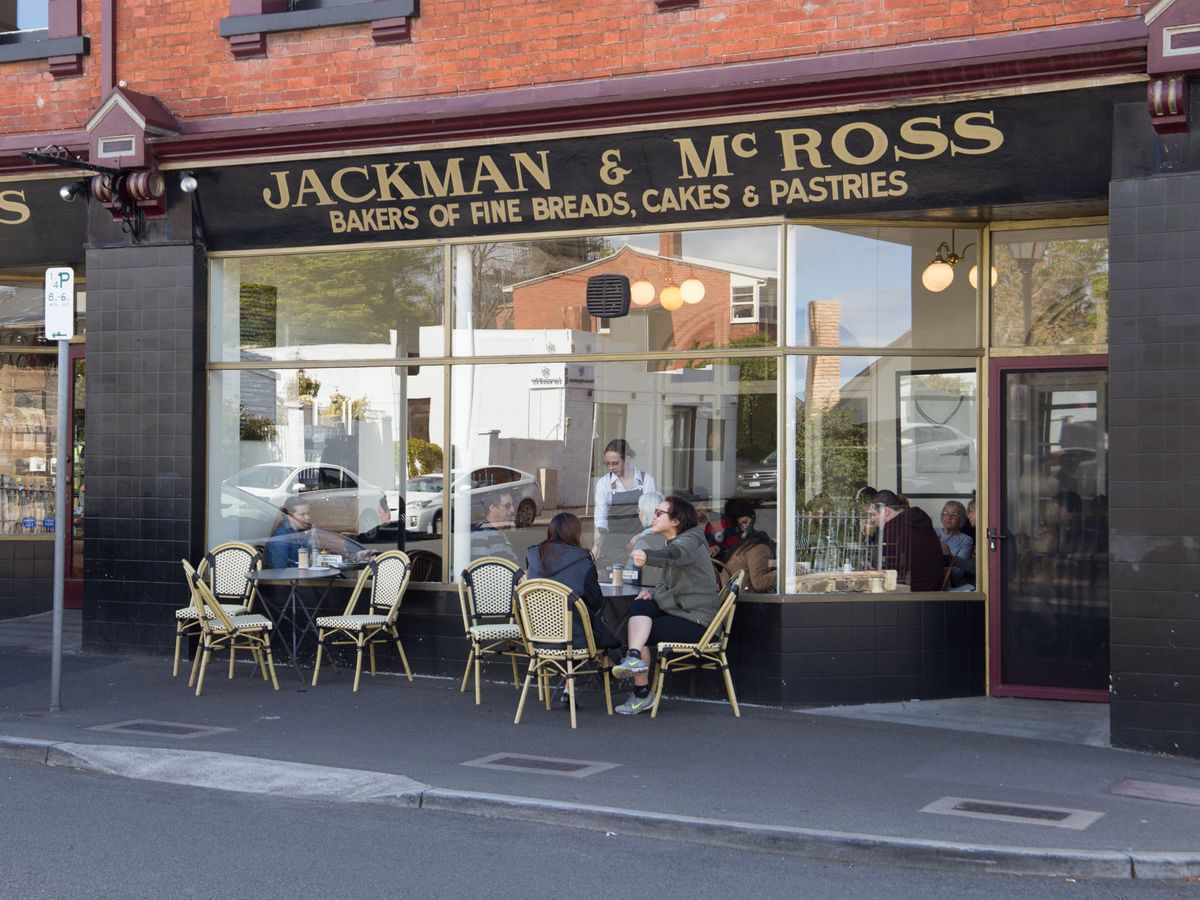 The old-style sign of Jackman &amp; McRoss bakery on a red brick building with huge windows into the dining room, and diners seated at outdoor patio tables.