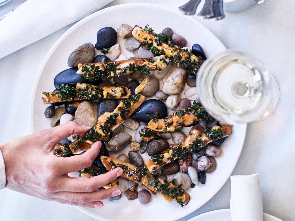 A big white plate of razor clams sitting on an elegant white tablecloth with a glass of white wine next to it