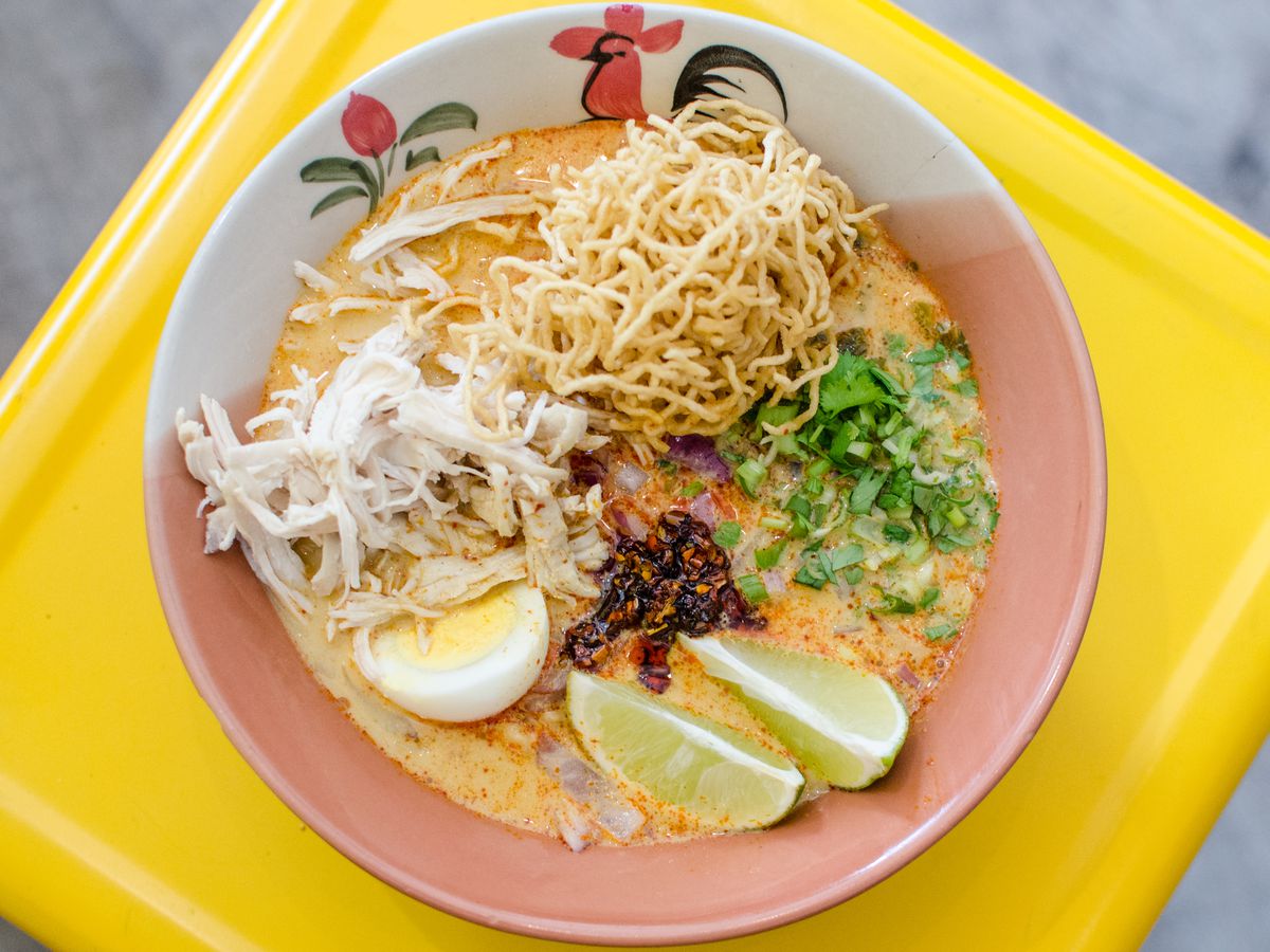 Overhead shot of khao soi on a bright yellow surface. The bowl has a traditional Thai pattern on it, including a rooster. The khao soi includes pickled mustard greens, an egg, lime wedges, a nest of crispy noodles, and more.