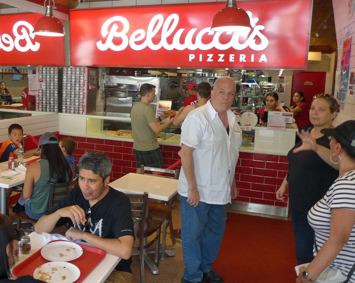 A man stands in his pizzeria with customers around him. 