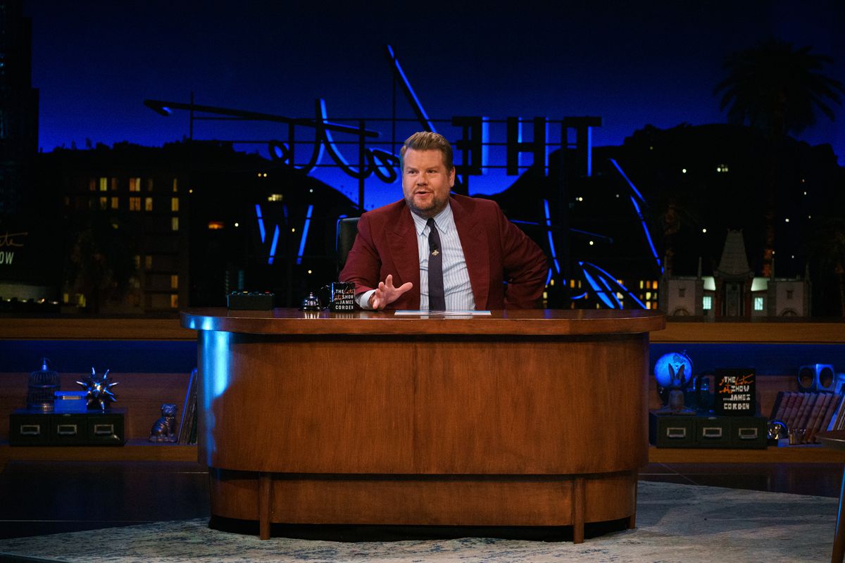 A celebrity, James Corden, sits behind a wooden desk on a late-night talk show host.