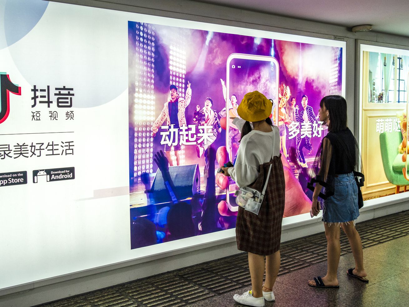 Two young women stand in front of a large digital billboard watching it display a Chinese ad for TikTok.