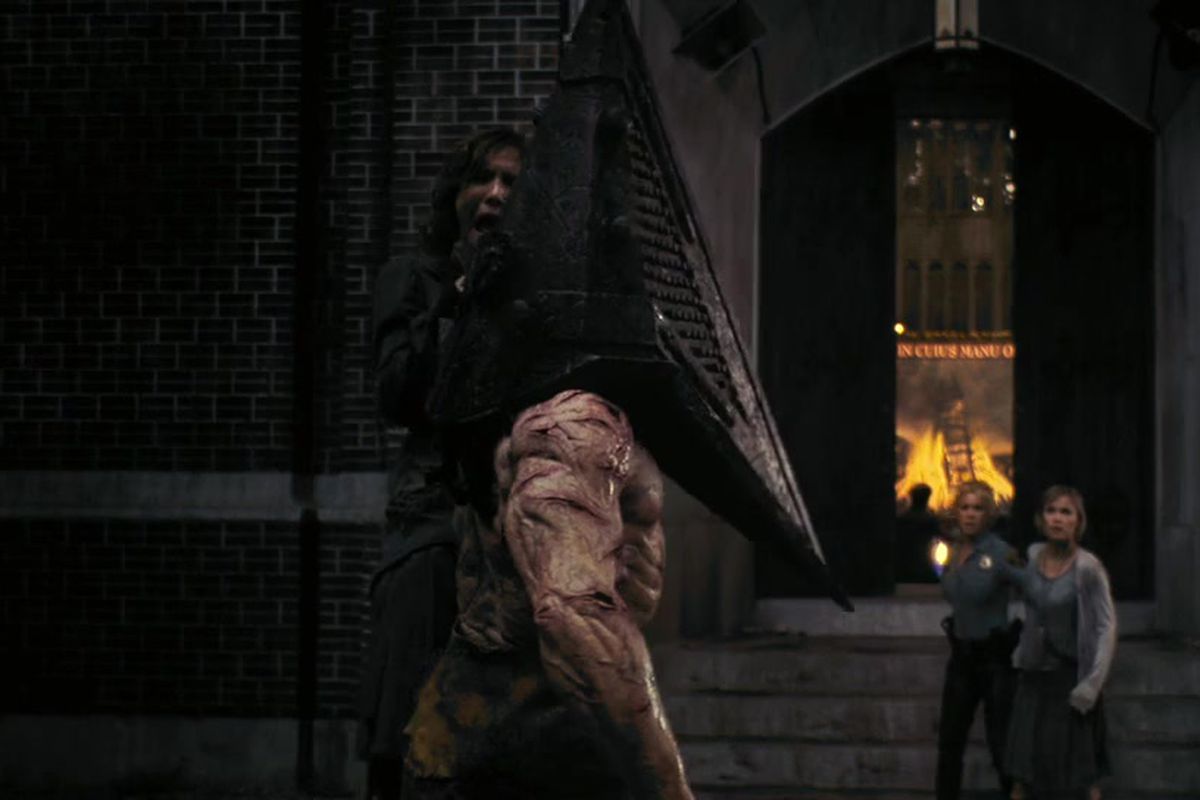 Pyramid Head (who has a pyramid for a head) from the Silent Hill movie stands in front of a church while holding a woman by the throat