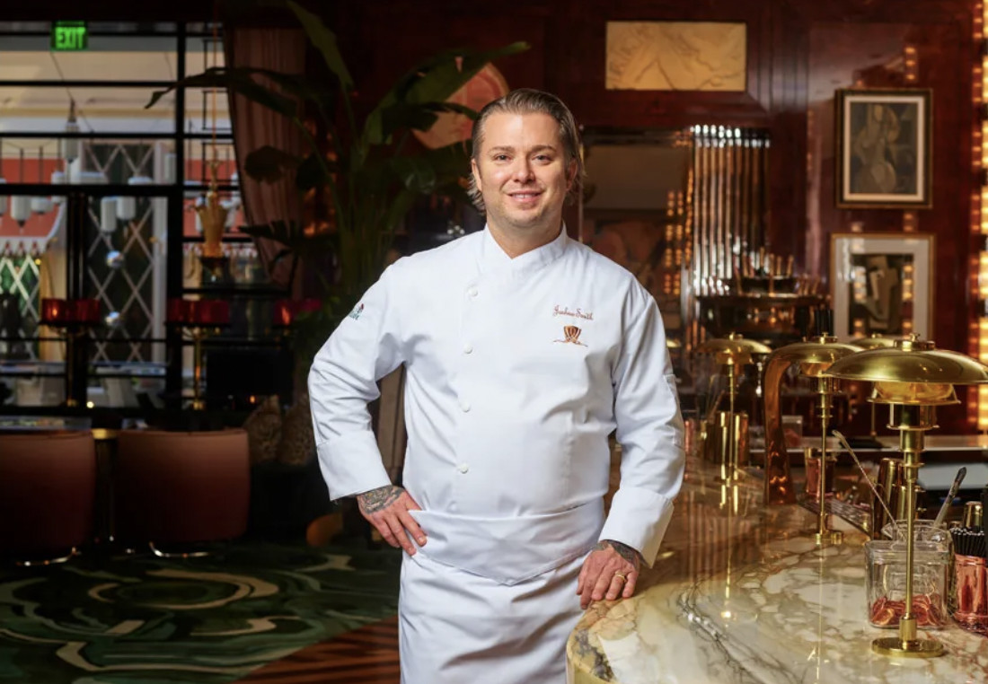 A man in a white chef’s jacket stands next to a decadent table. 