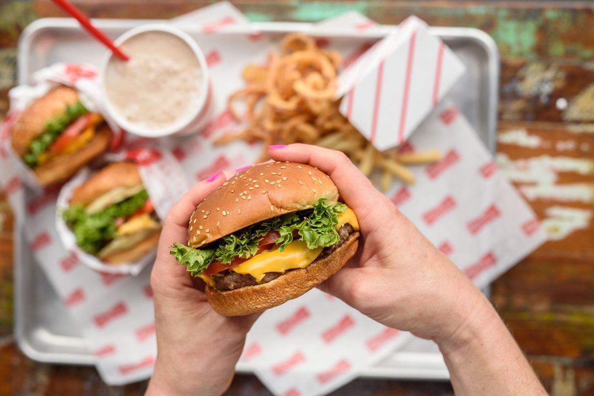 Hands hold onto a cheeseburger from Tasty Burger, held aloft above more burgers and onion rings