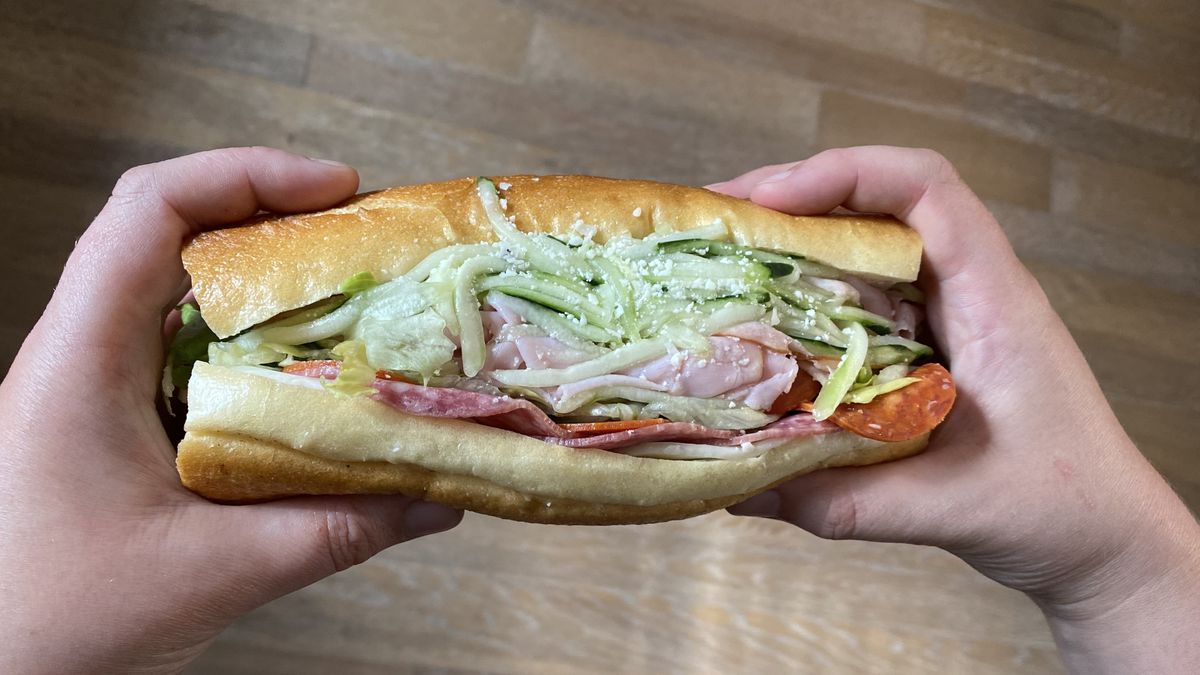 Two hands hold a sandwich filled with ham, pepperoni, salami, sliced cucumbers, and parmesan from Taste Tickler in Portland, Oregon.
