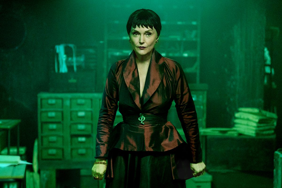 Miranda Richardson wears a tightly fitted red blazer and black pencil skirt combo, with her hair in a choppy black bowlcut. She stands in a green-lit dark office. 