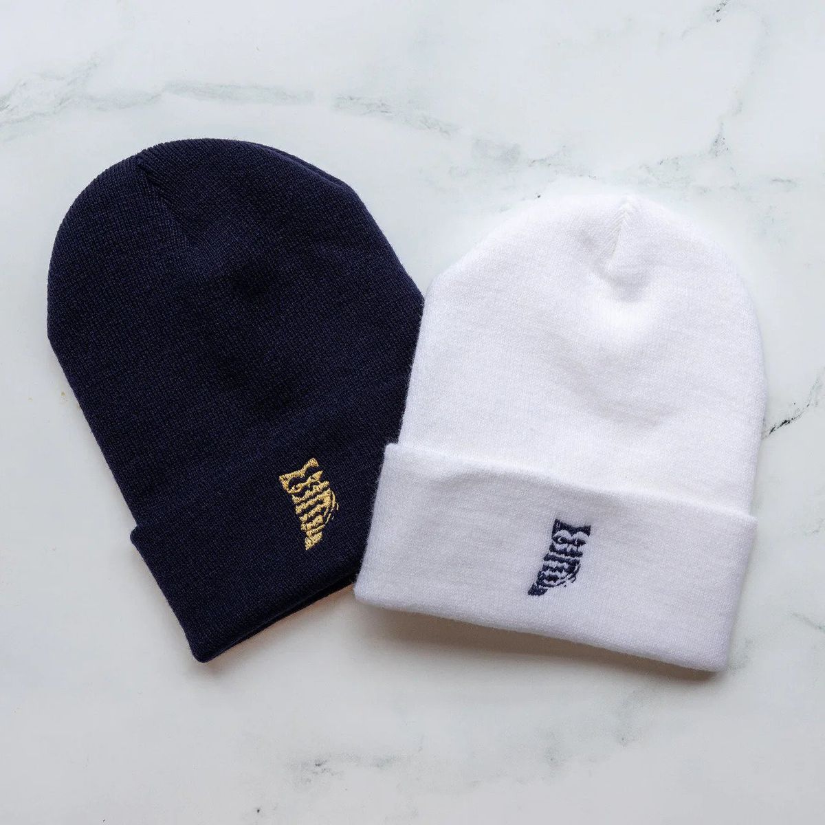 Two knit beanies, one navy, one white, both with an embroidered owl logo