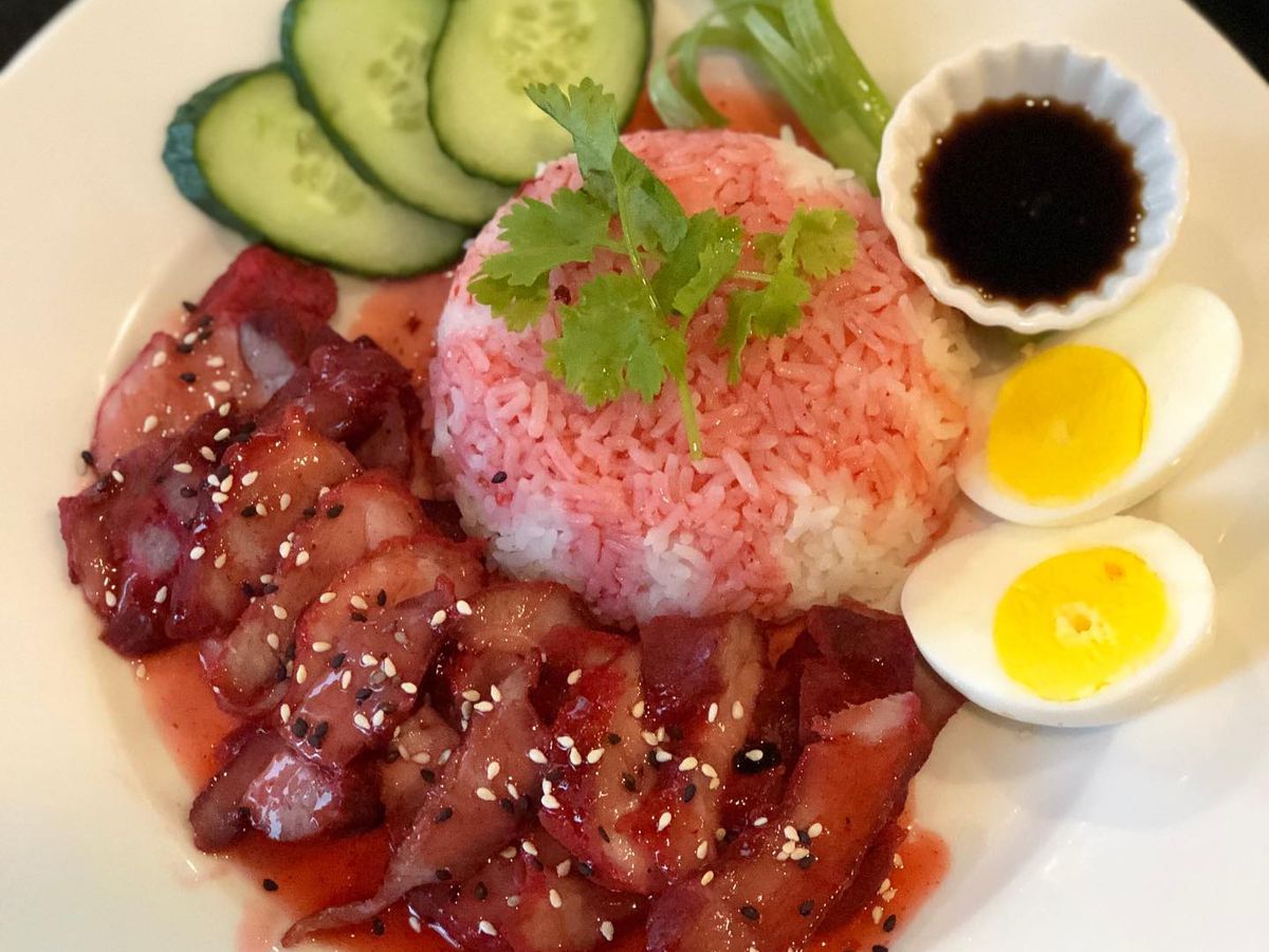 A plate shows strips of meat sprinkled with sesame seeds next to a mound of rice, a halved boiled egg, cucumbers, and a dark sauce. 