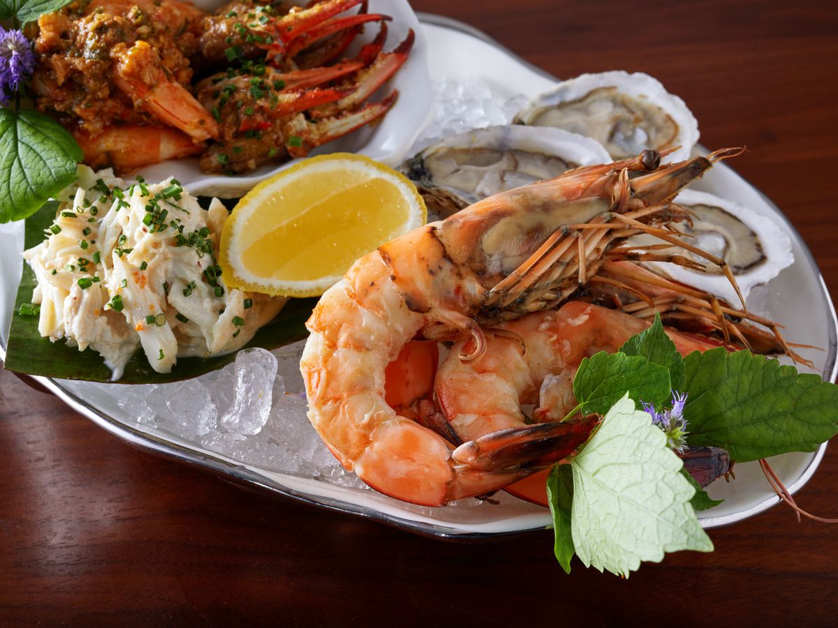 A silver platter topped with crushed ice holding a mix of large steamed prawns, crab claws, oysters on the half shell, and smoked fish dip.