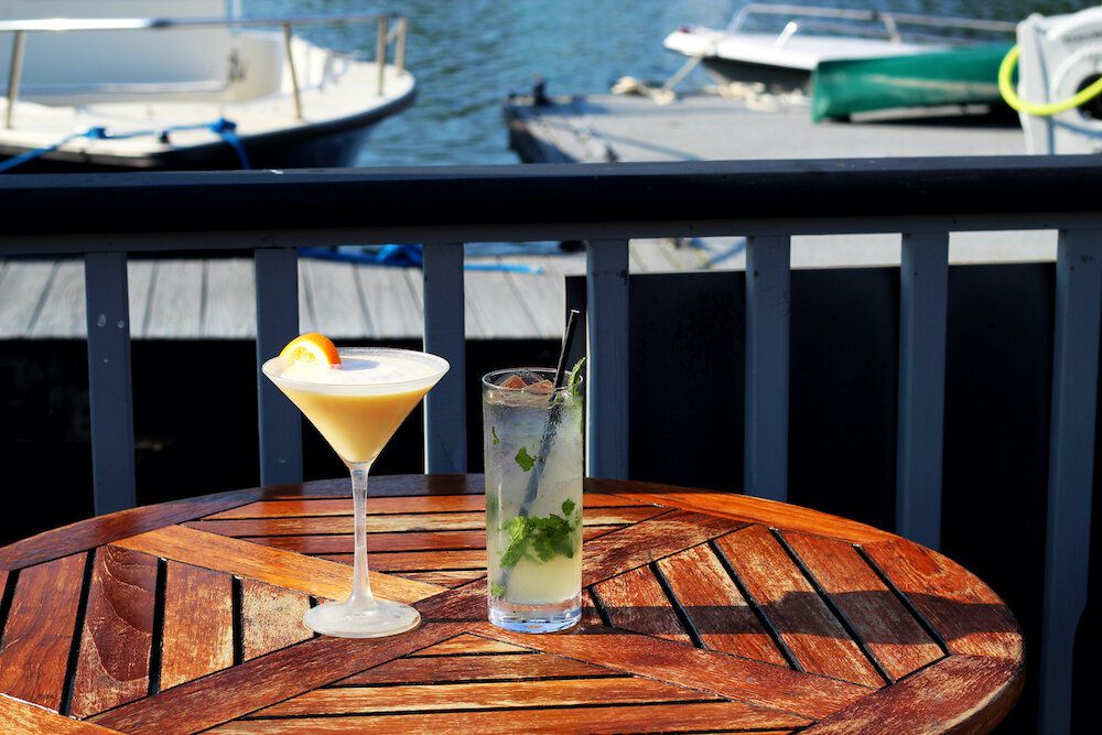 Two cocktails sit on a wooden table in an outdoor dining area, with boats and water visible in the background.