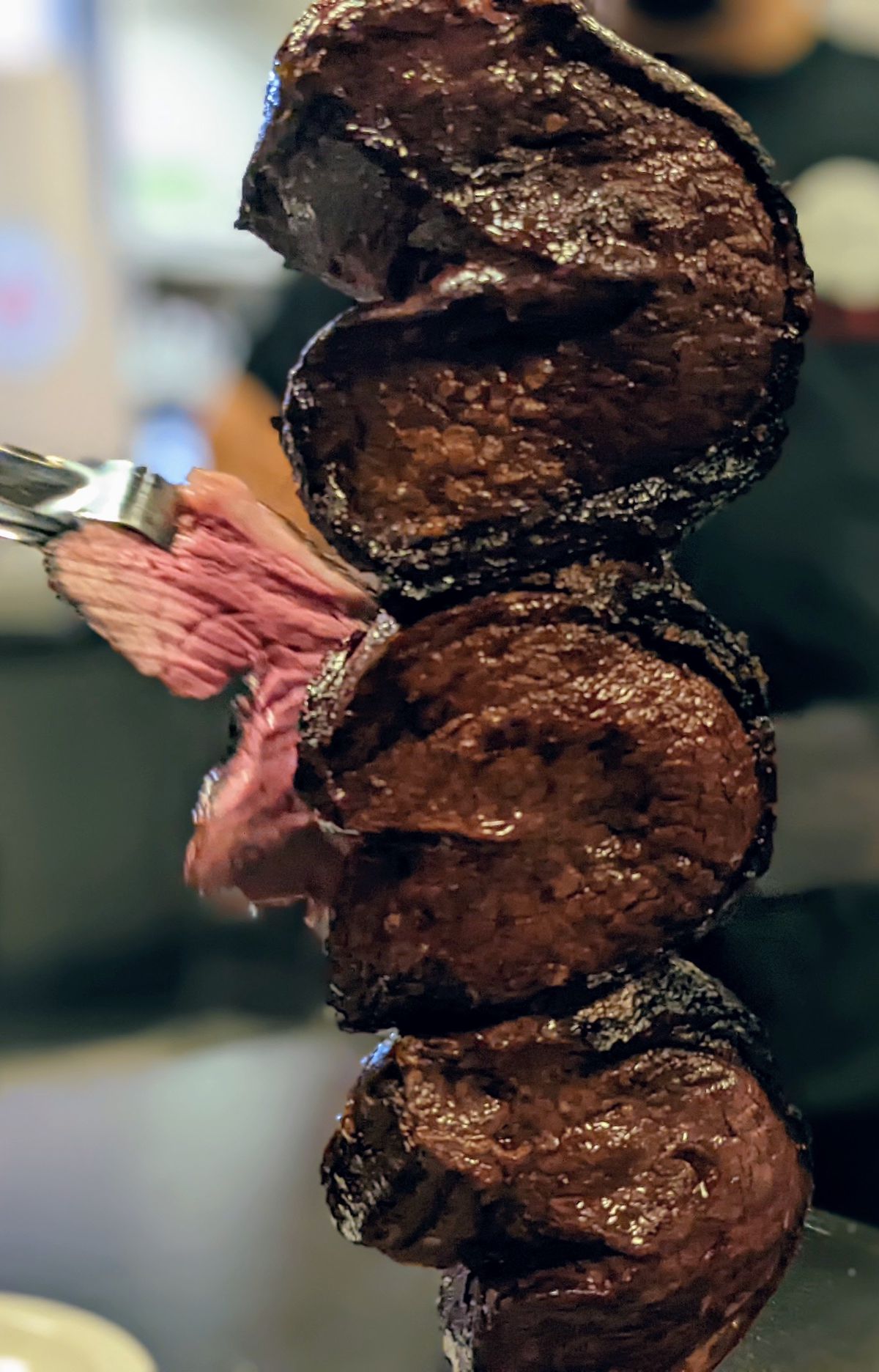 A pair of tongs pulls out a slice of rare meat from a skewer.
