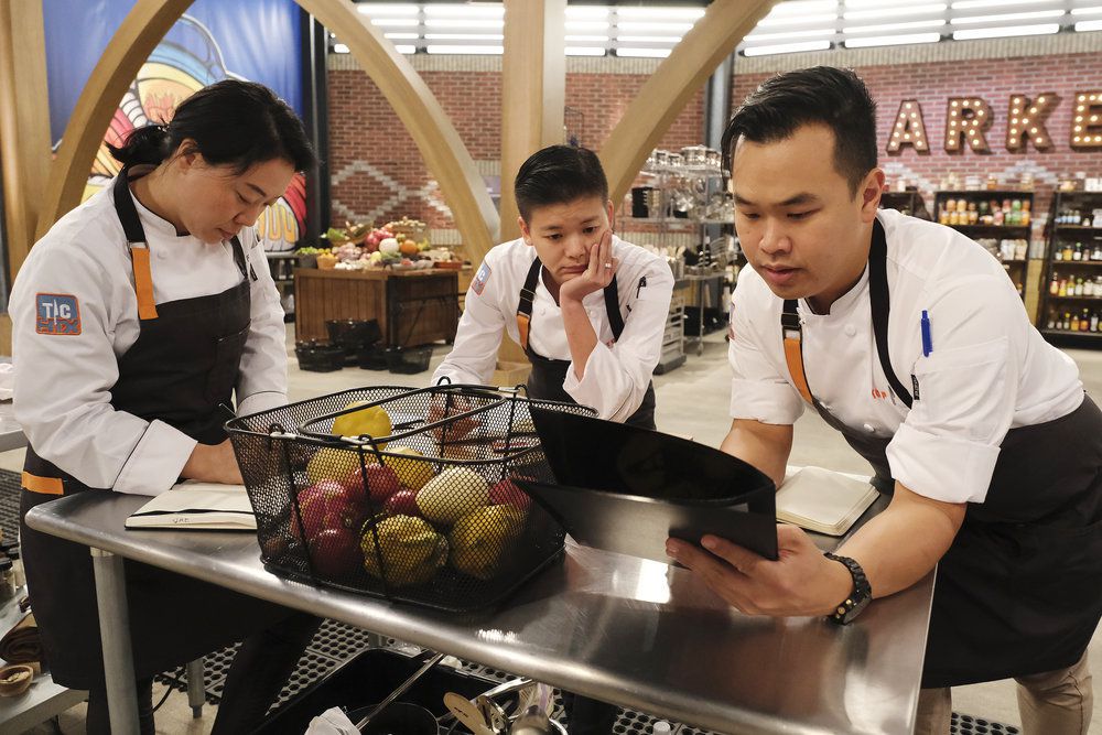 Three chefs standing at a metal table looking at folders of information.