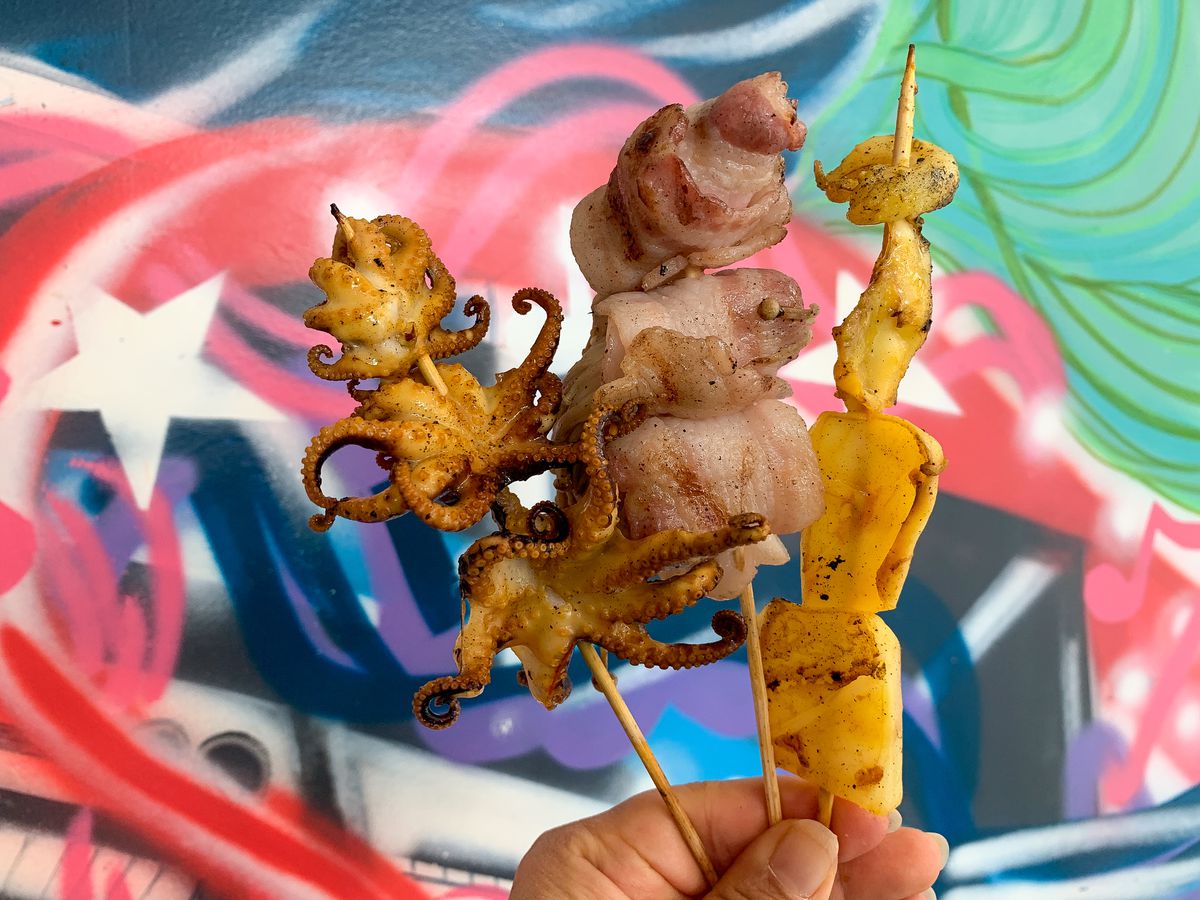 A hand holding up three skewers against a colorful painted wall.