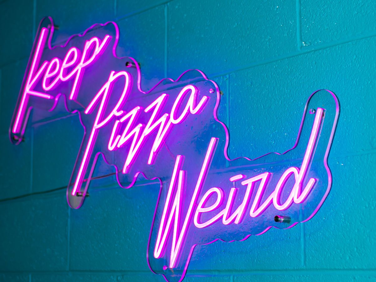 A bright pink neon sign lit up against an aqua, cinderblock wall. In cursive, the neon sign says, “Keep Pizza Weird.”