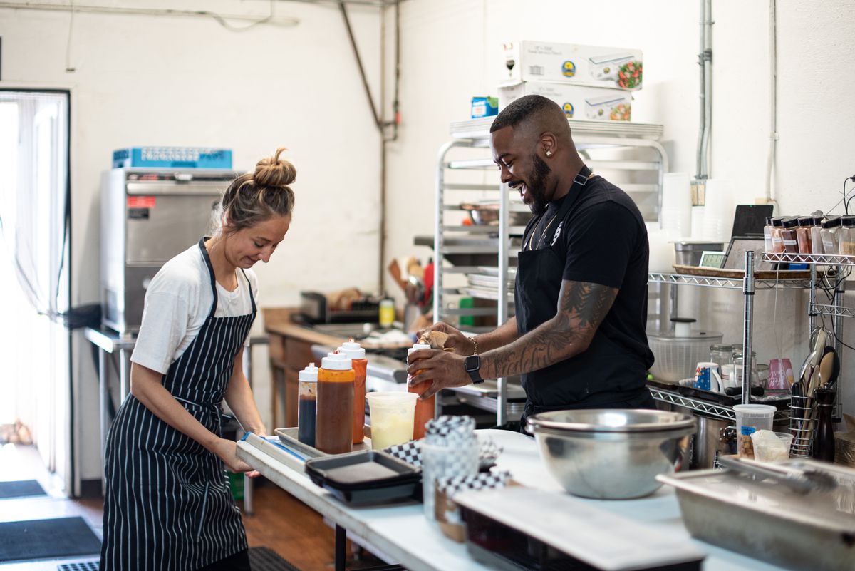 A female chef and a male chef work together over a table to make chicken wings.