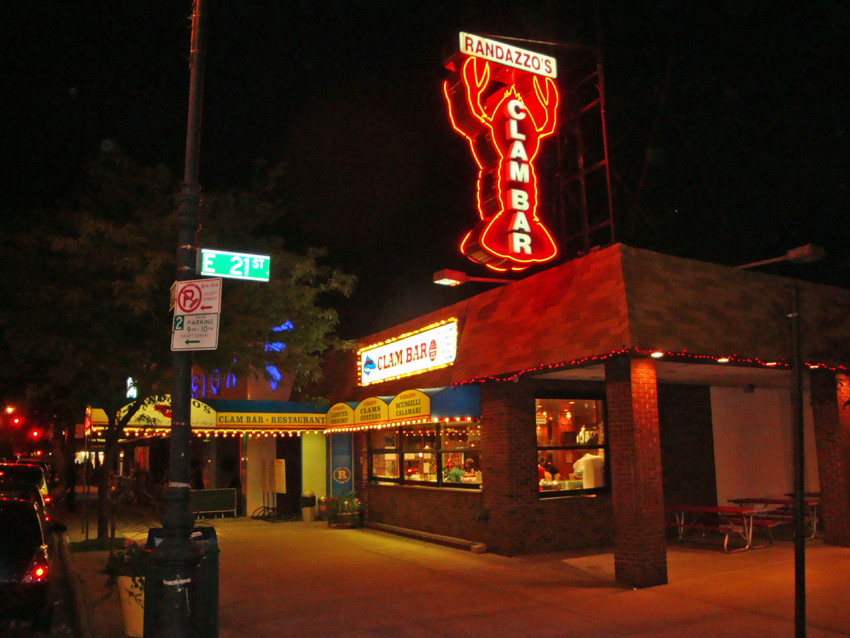 A neon sign in the shape of a lobster with the words “Randazzo’s Clam Bar” gleams above a restaurant at night