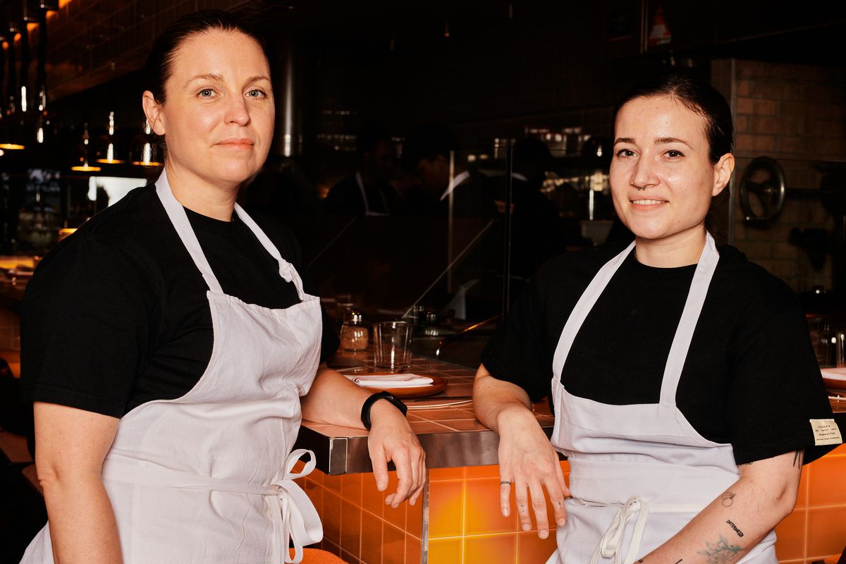 Chef Melissa Rodriguez and pastry chef Georgia Wodder stand for a portrait, both clad in black shirts and cream-colored aprons