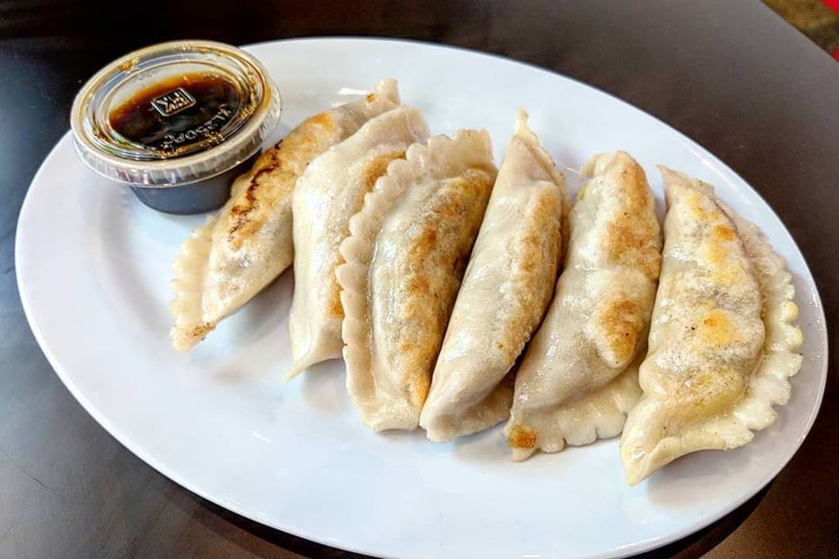 Six pan-seared dumplings sit on a white plate. A plastic cup of a soy-based dipping sauce is next to them.