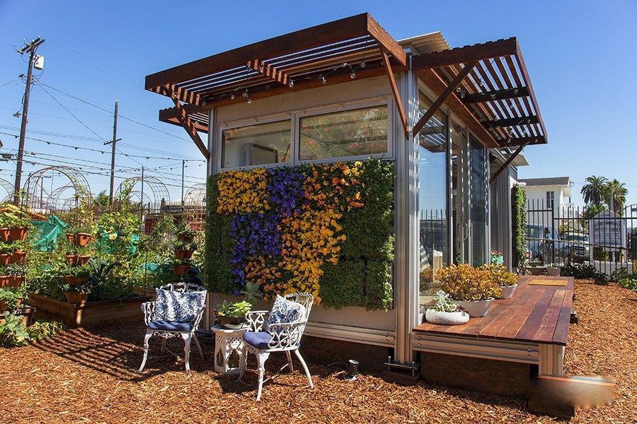 A tiny home with a wall of plants and flowers on the side of it. Two chairs and table are next to the flower wall. 