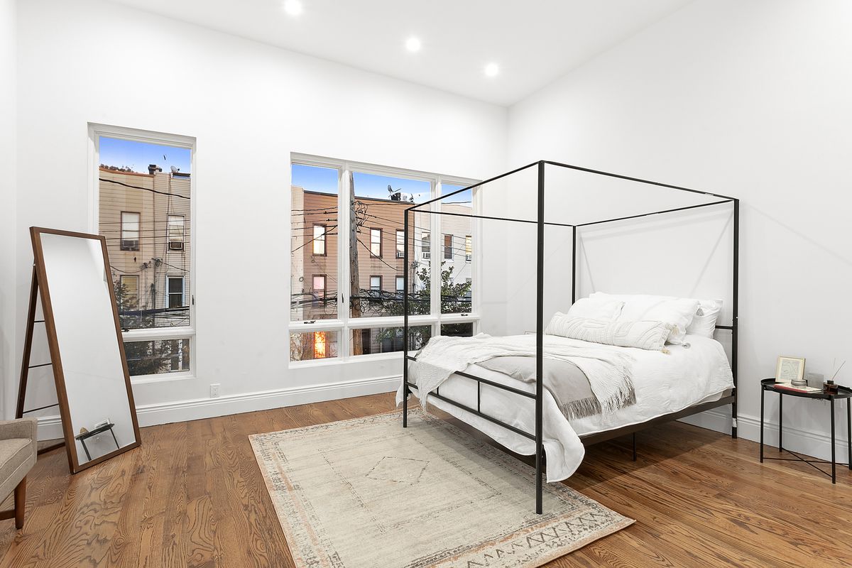 A bedroom with large windows, hardwood floors, a standing mirror, and a medium-sized bed.
