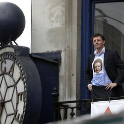 An office worker wearing a depiction of former British Prime Minister Margaret Thatcher on his shirt watches people protest along the ceremonial funeral procession for the former prime minister from an office balcony in central London, Wednesday, April 17, 2013.