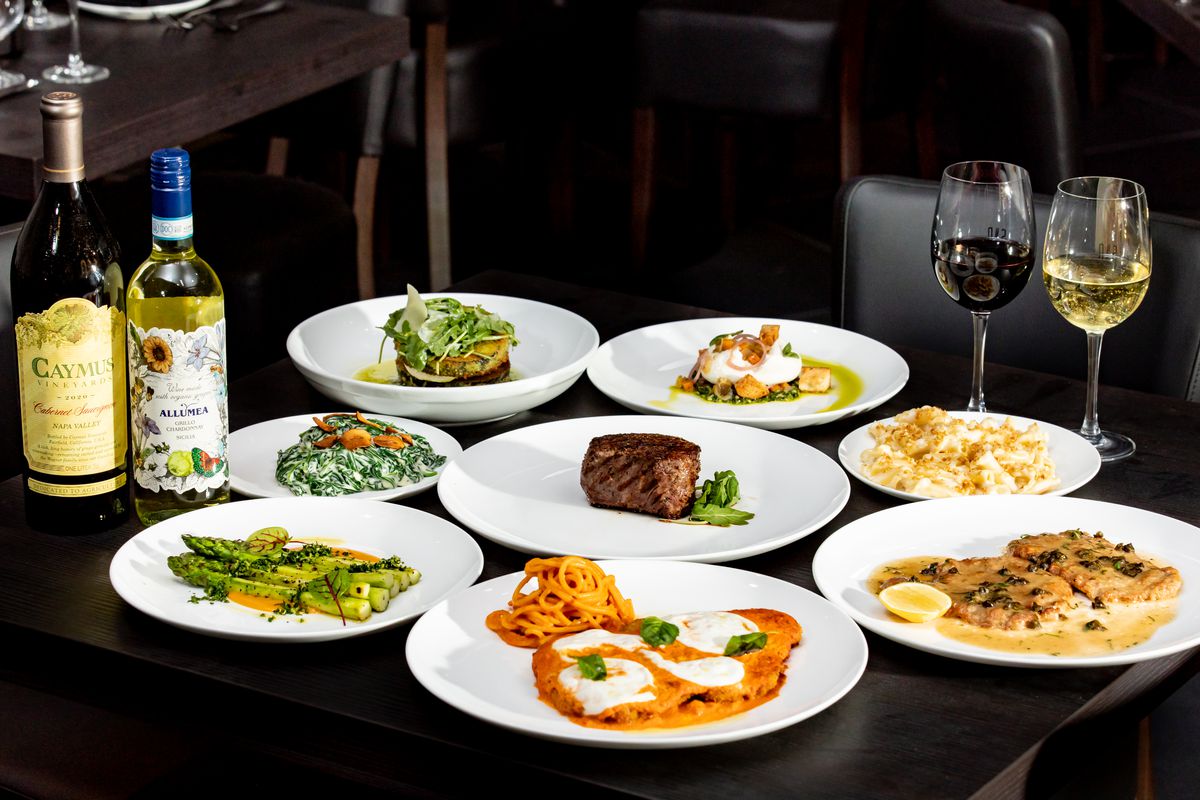 A spread of plated Italian dinner entrées on a table in a restaurant alongside bottles of wine.