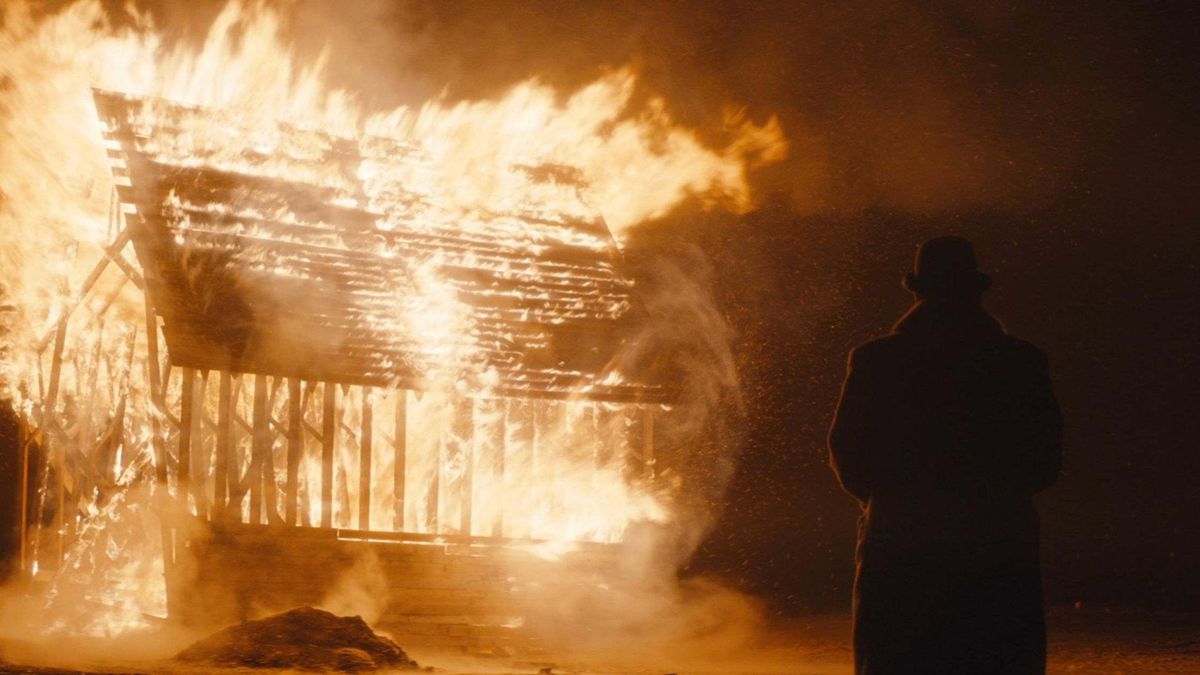 A figure in a large winter coat and a bowler cap stands in front of a building set on fire in the middle of the night.