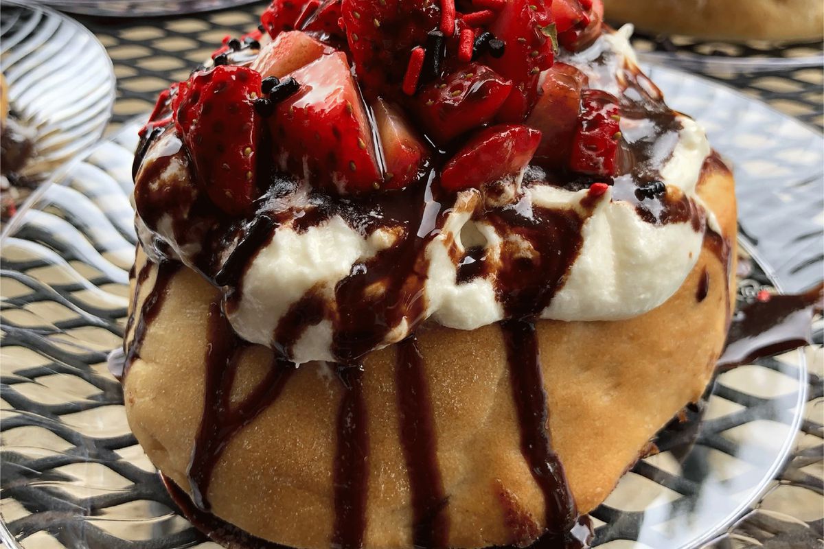 A cinnamon roll topped with chocolate sauce, white icing, and strawberries 