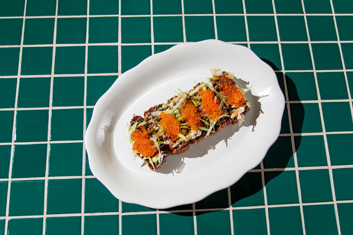 A green tiled countertop with a white dish and five rectangular latkes topped with scallions and orange trout roe.