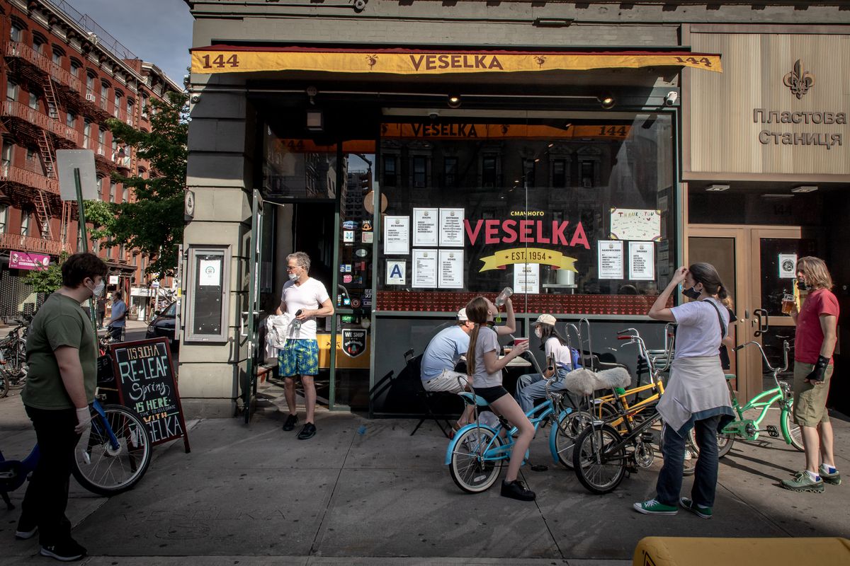 People gather on bikes outside Veselka while a patron in a white t-shirt and blue patterned shorts walks out; most are wearing face masks.