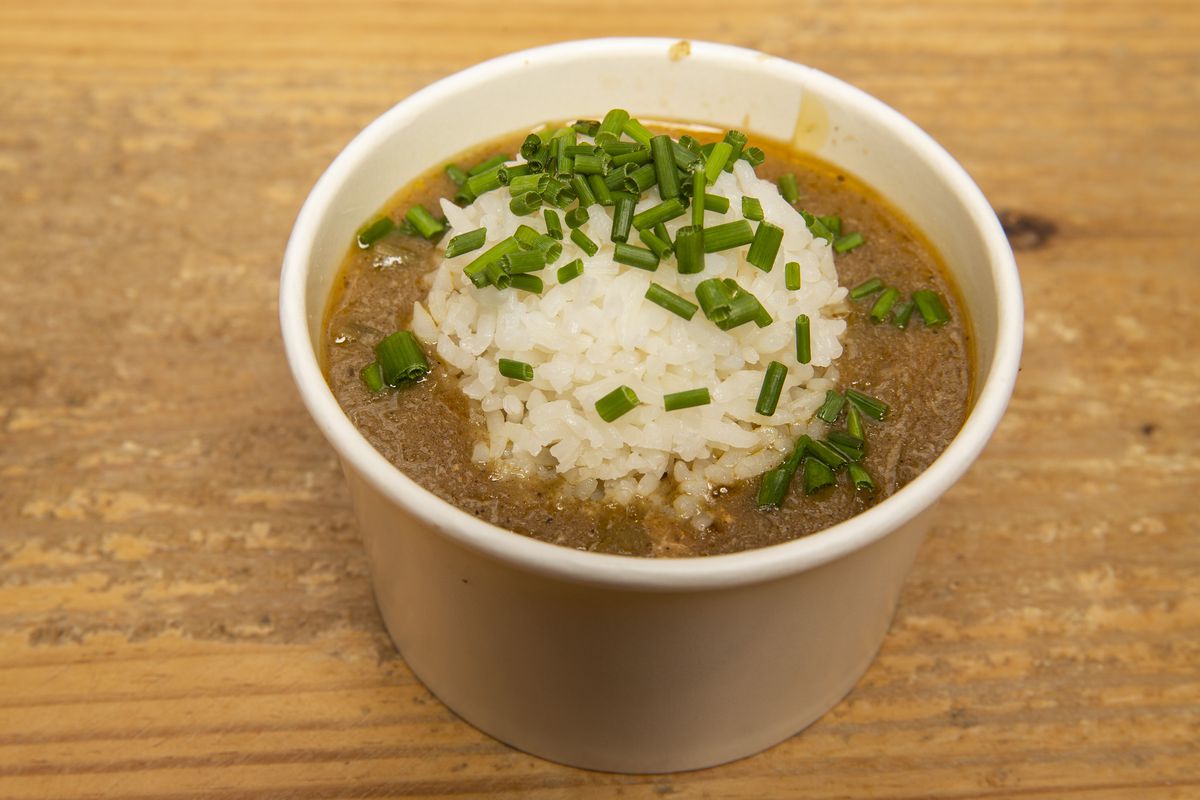 A white paper container holds a serving of chicken gumbo topped with a pile of white rice and chopped chives.