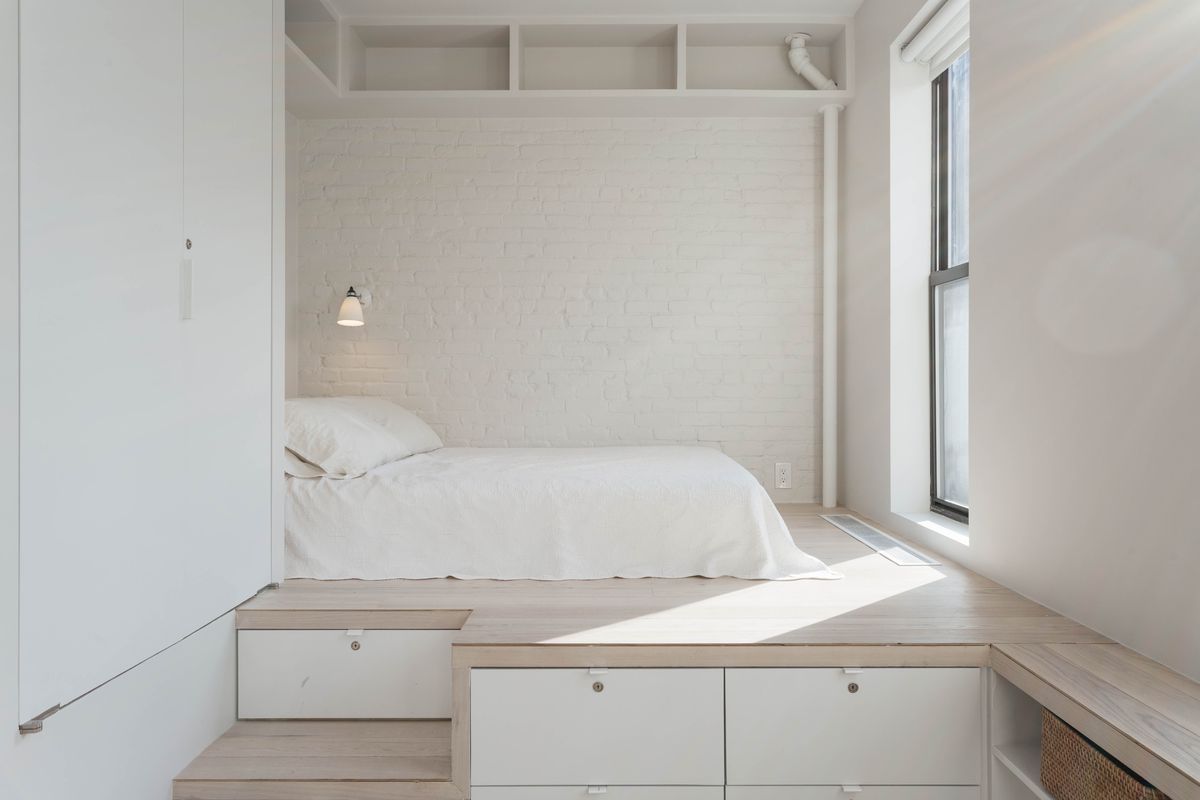 An alcove in a studio with a small bed, white exposed brick, pale oak floors, and a window.
