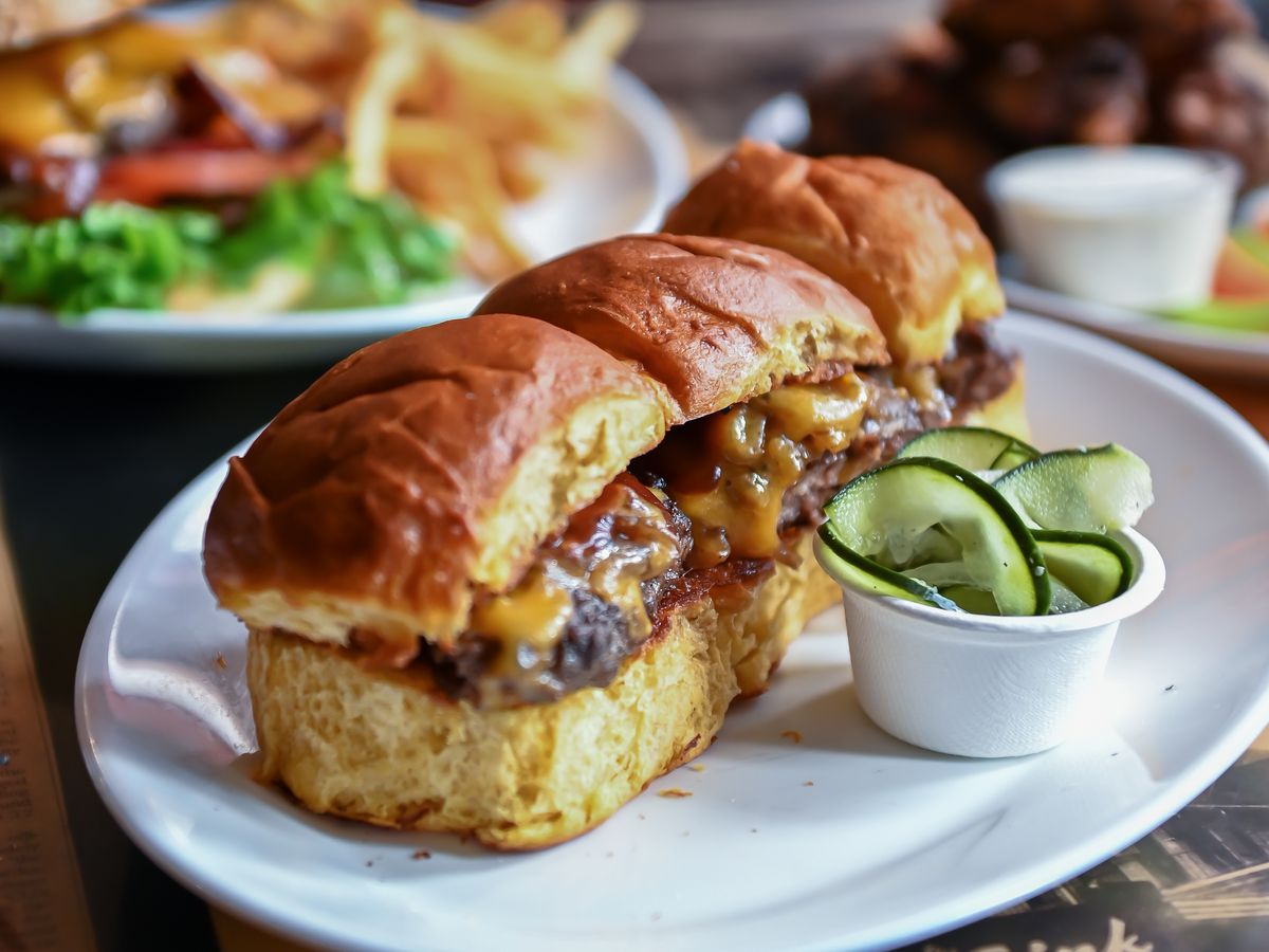 Three cheeseburger sliders on a plate with a small cup of pickles.