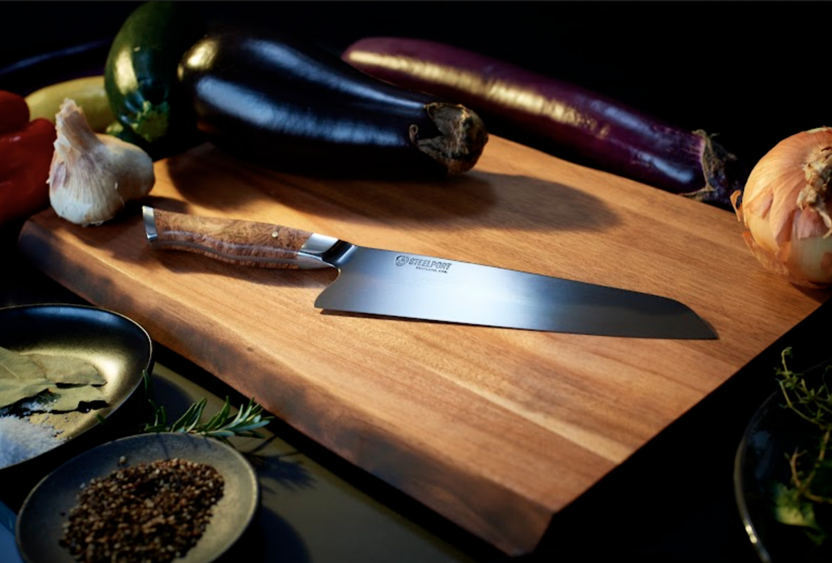 Steelport chef’s knife on a cutting board, sitting next to produce.