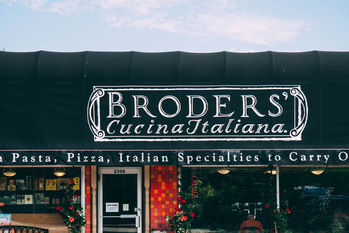 The exterior of Broders’ Cucina Italiana: A black awning with white writing and a brick storefront with windows, against a blue sky.