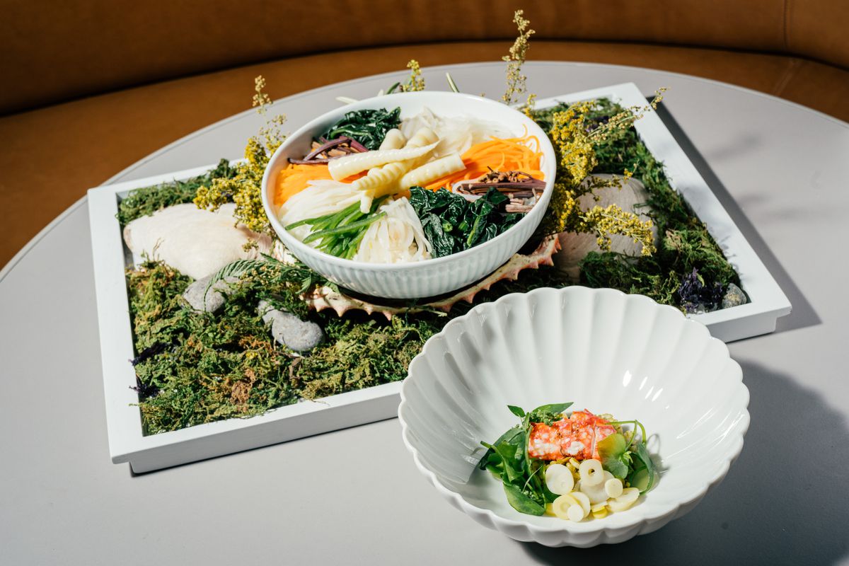Two white bowls, one with vegetables and another with small slices of king crab, are arranged on a white table.