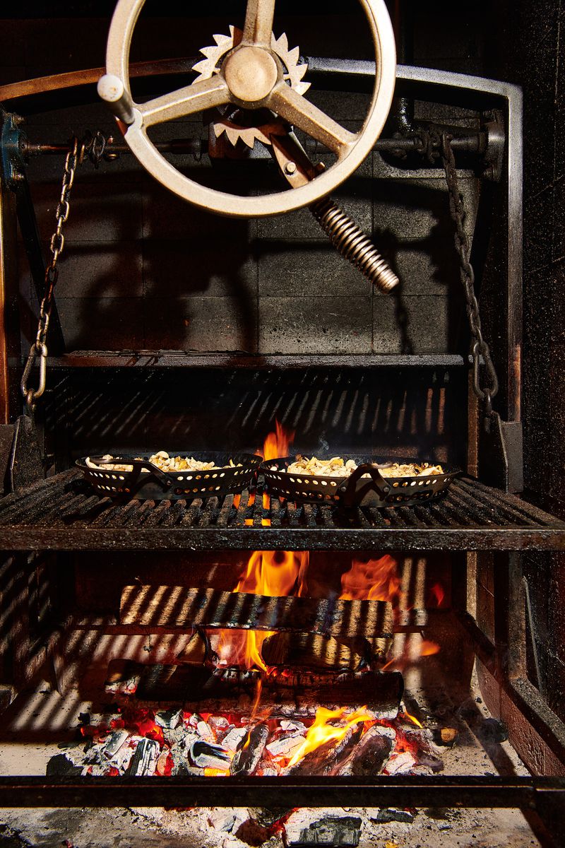 A large round crank controls a large grill set over a wood fire. 