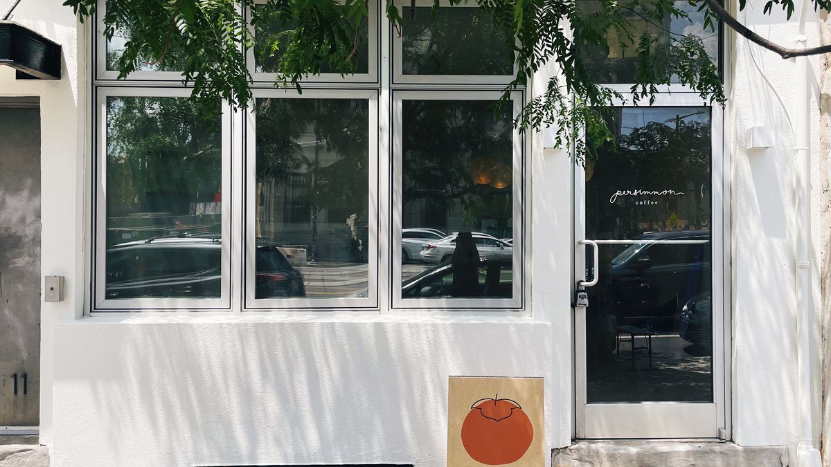 The outside of a shop with a white facade with a green tree and a sandwich board with an orange persimmon that reads Coffee.