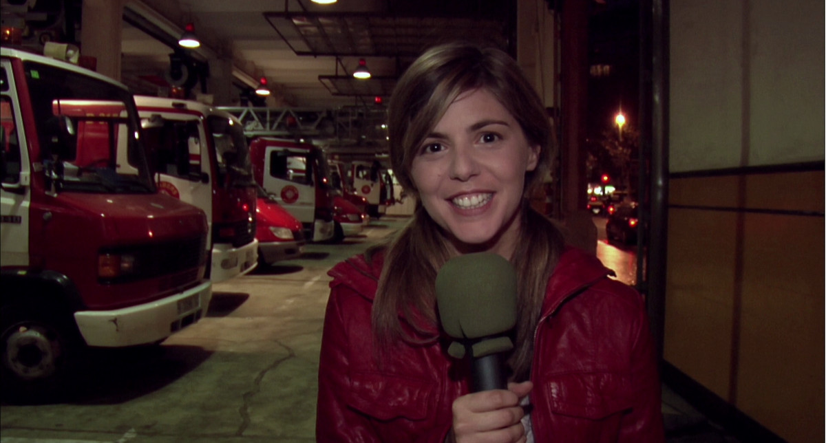 Una mujer joven con una chaqueta roja habla por un micrófono en una transmisión de televisión desde una estación de bomberos en Rec.