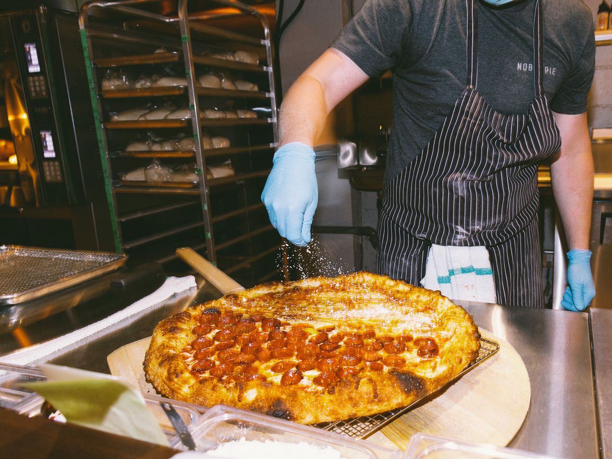 A chef in gloved hands sprinkles cheese on top of a pizza in a restaurant kitchen