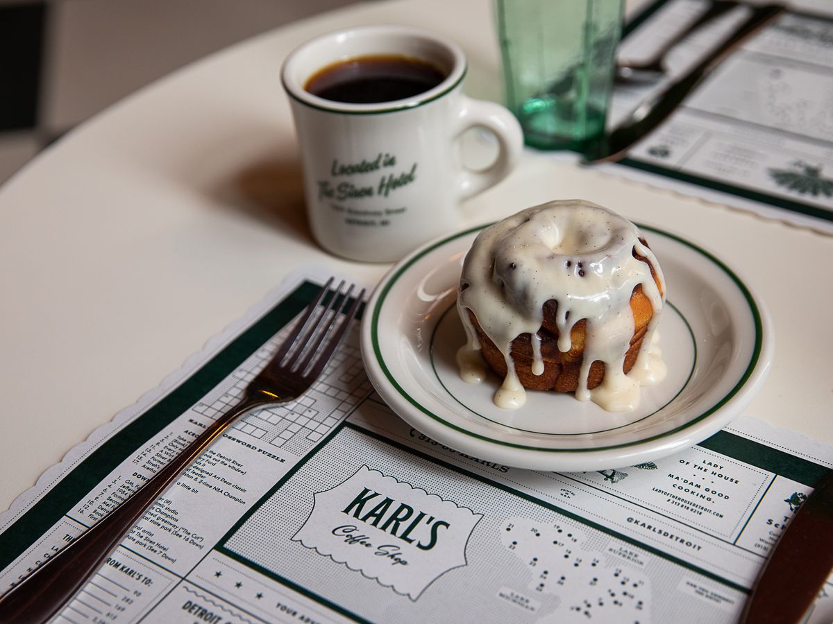 A cinnamon roll dripping with thick, white icing is sitting on a small green and white plate next to a branded coffee mug and on top of a Karl’s Coffee Shop placemat.